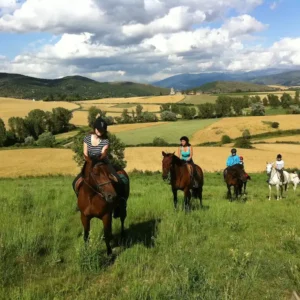 Grupo de chicas montando a caballo en fila en un paisaje verde con campos de cultivo de fondo.